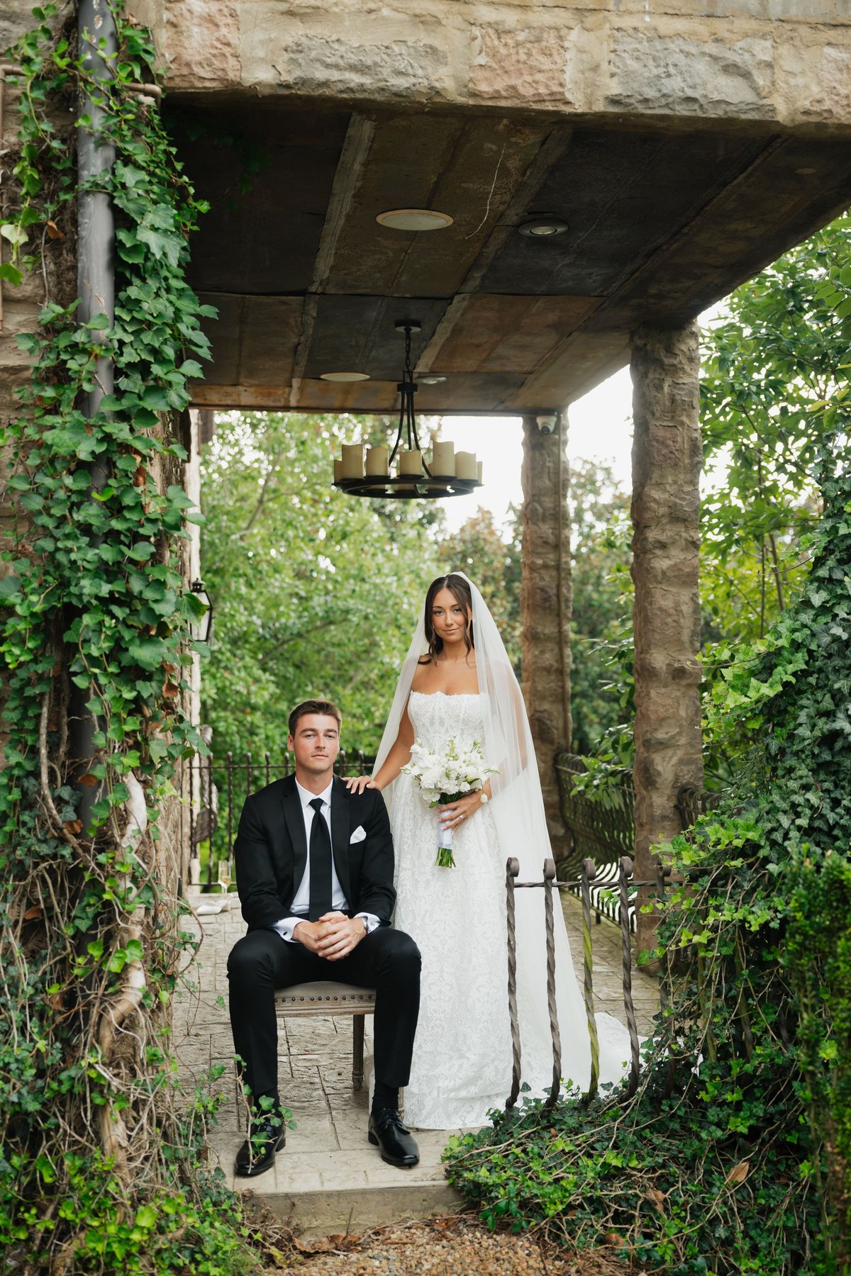 Bride standing beside her seated groom under an ivy-covered stone pavilion