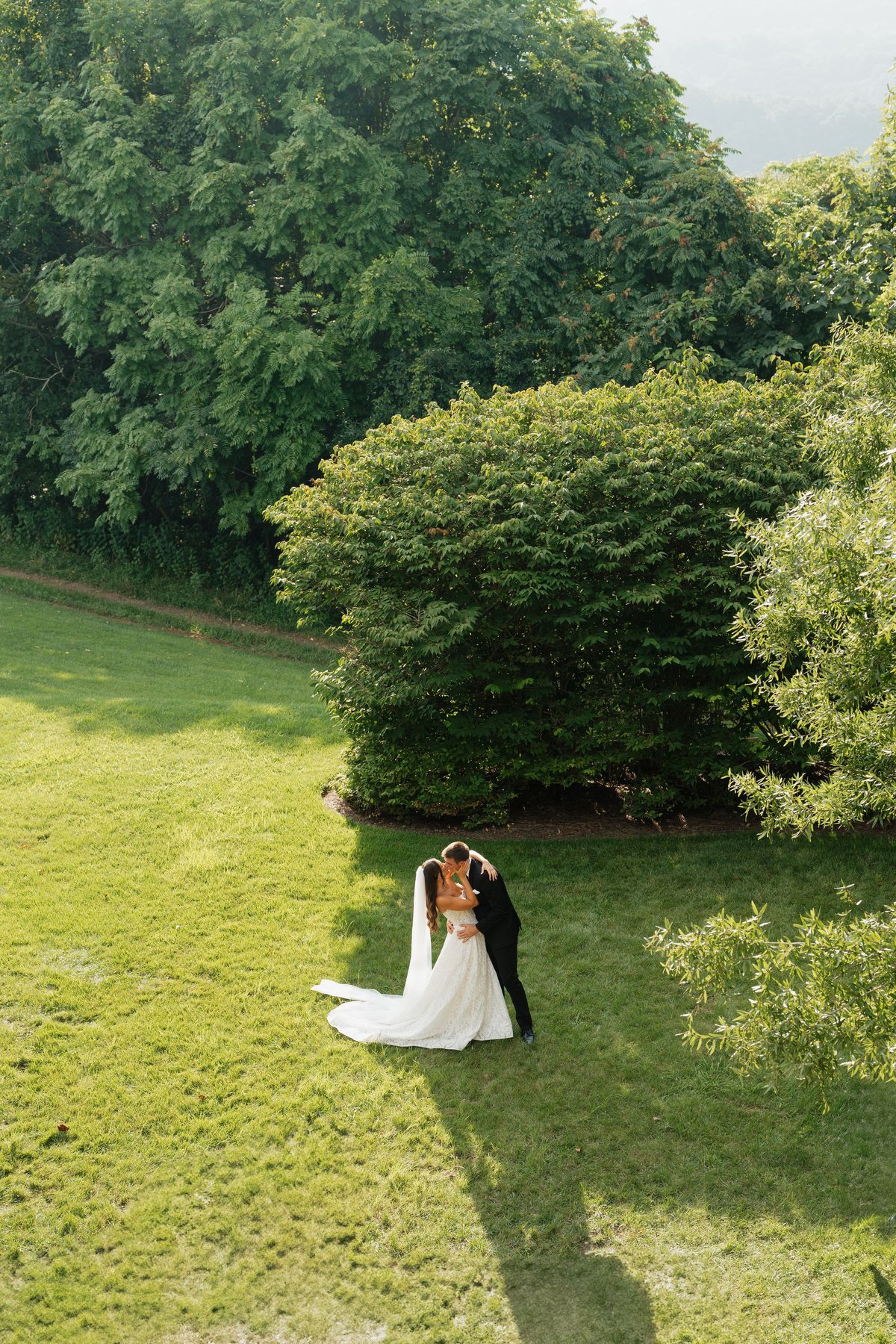Bride and groom embracing on a wide green lawn framed by summer trees
