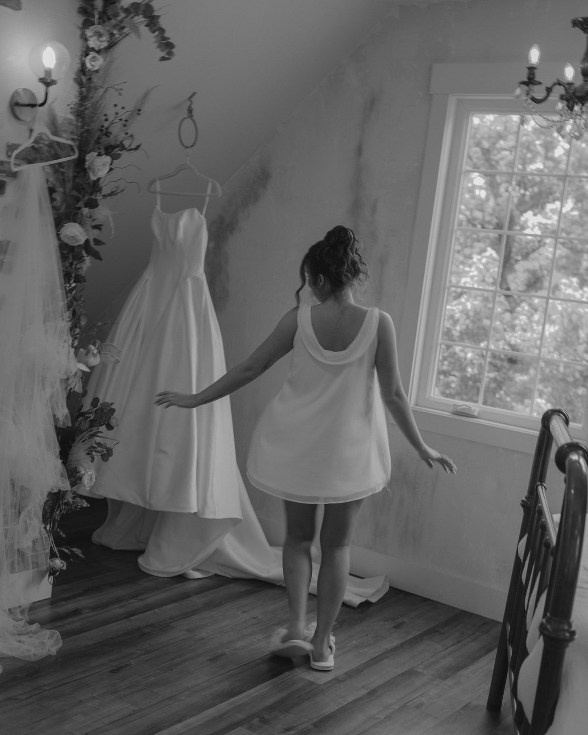 Bride reaching toward her wedding dress hanging in a sunlit attic room