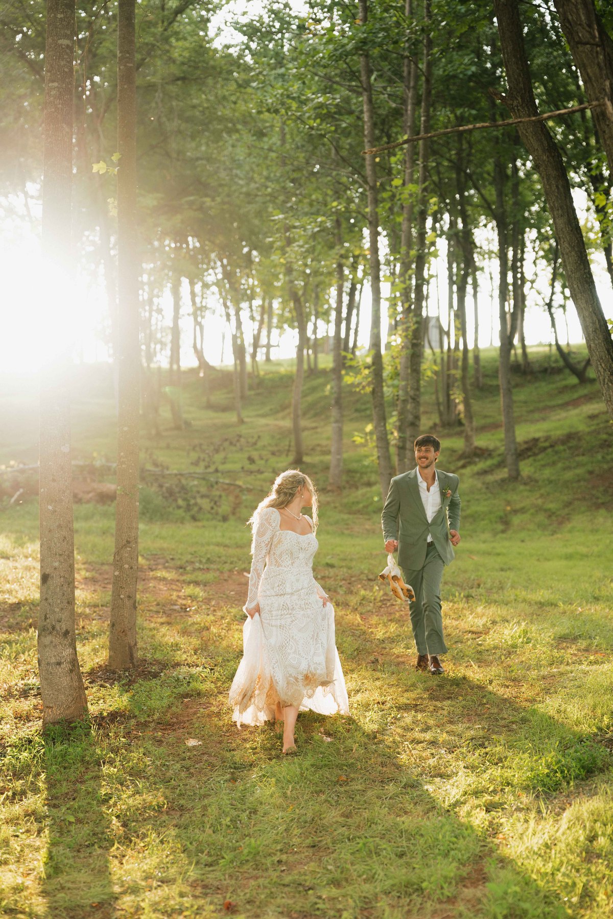 Bride and groom walking through a sunlit forest, groom in a sage green suit